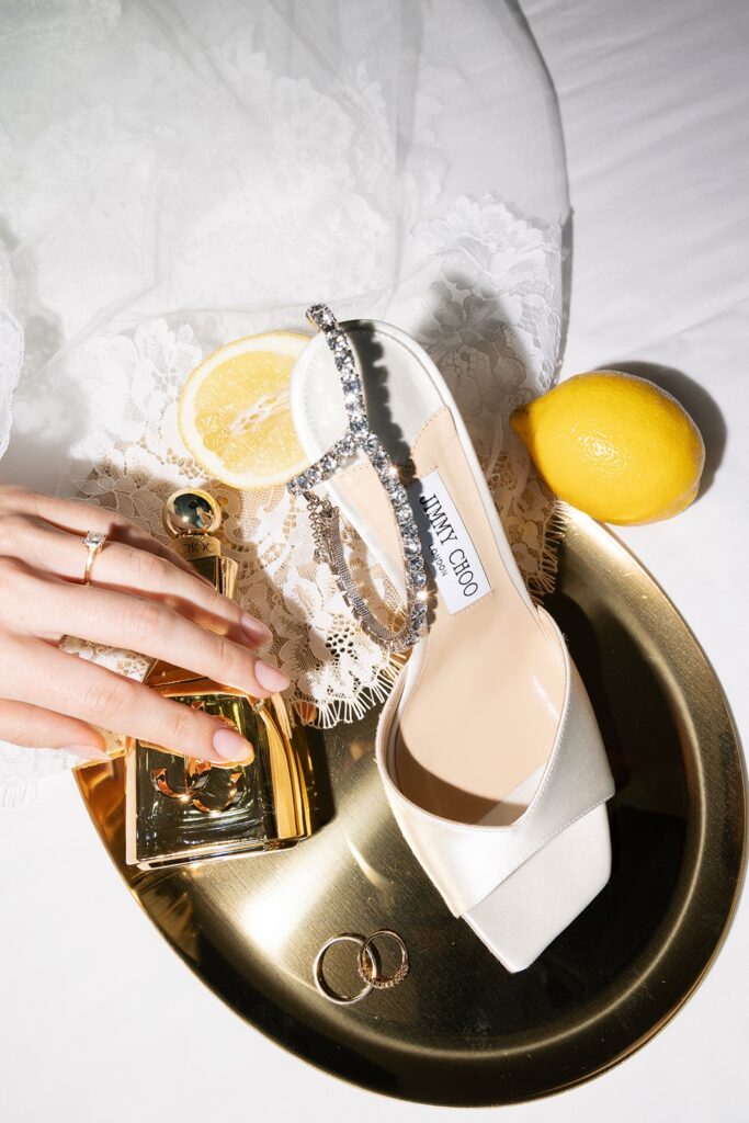Elegante Jimmy Choo Brautschuhe mit funkelnden Riemchen auf goldenem Tablett, daneben Chanel Parfum, Eheringe und Zitronen als sommerliches Detail
